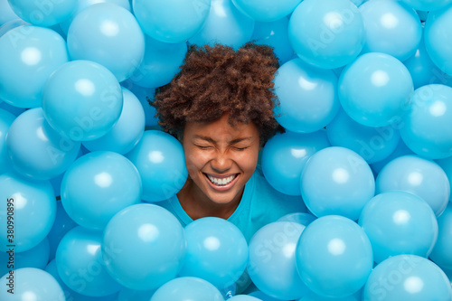 Overjoyed curly haired Afro American woman feels amused and entertained on party has fun and sticks out head through wall decorated with blue balloons expresses happy emotions. Festive event