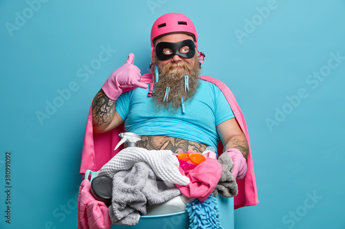 Bearded man makes call me gesture dressed in superhero costume entertains children and does laundry at same time poses against blue background. Husband or dad does housework washing at home.