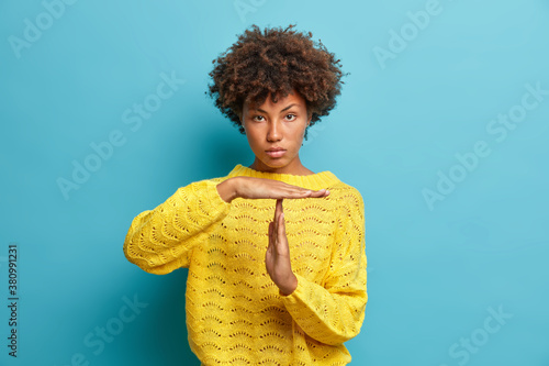 Serious self confident curly haired Afro American woman makes timeout gesture demonstrates limit asks to stop dressed in yellow knitted jumper isolated on blue background. Body language concept photo