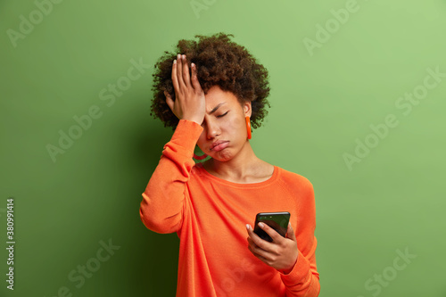 Upset depressed Afro American woman makes face palm as forgot about something important holds mobile phone expresses sorrow and regret blames herself isolated on green background. Negative emotions