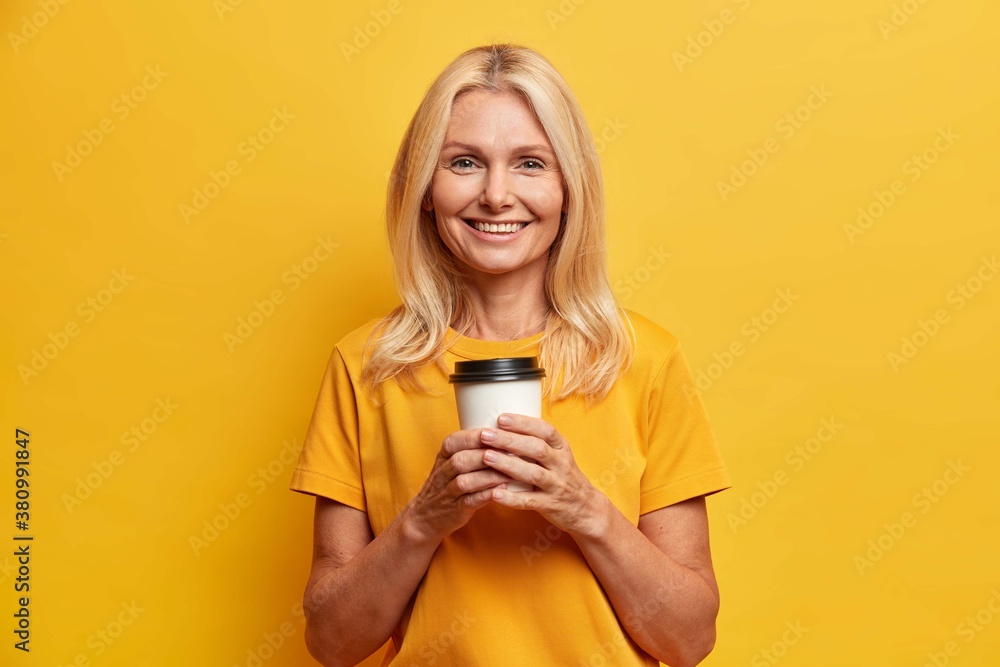 Horizontal shot of pretty wrinkled middle aged woman holds disposable cup of coffee enjoys free time and looks happily at camera dressed in casual t shirt isolated on vivid yellow background Horizontal shot of pretty wrinkled middle aged woman holds disposable cup of coffee enjoys free time and looks happily at camera dressed in casual t shirt isolated on vivid yellow background