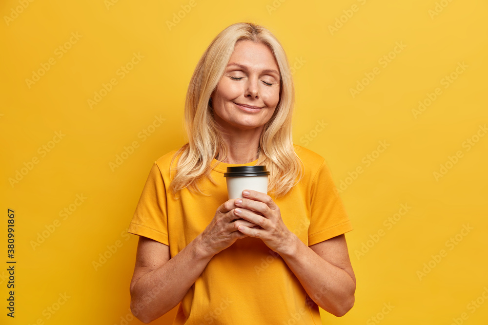 Photo of relaxed satisfied woman stands with closed eyes holds paper cup of coffee has dreamy peaceful expression smells aromatic beverage wears casual t shirt isolated on yellow background. Photo of relaxed satisfied woman stands with closed eyes holds paper cup of coffee has dreamy peaceful expression smells aromatic beverage wears casual t shirt isolated on yellow background.