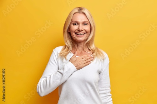 Happy grateful blonde woman in her fifties holds hand on chest and smiles broadly swears be loyal thanks for honesty smiles gladfully dressed in casual white jumper isolated on yellow background photo