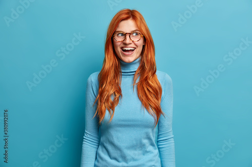Horizontal shot of overjoyed ginger young woman with European appearance laughs out loudly has fun and rejoices nice event wears optical glasses casual turtleneck isolated on blue background