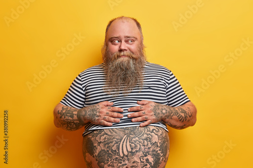 Overweight bearded man with fat belly and tattoos has no sport bad eating habits looks thoughtfully aside thinks about weight loss dressed in undersized striped shirt isolated on yellow background