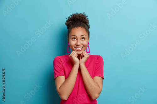 Joyful young Afro American woman keeps hands under chin concentrated above with enjoyment has dreamy expression dressed in casual crimson t shirt isolated on blue background. Human emotions.
