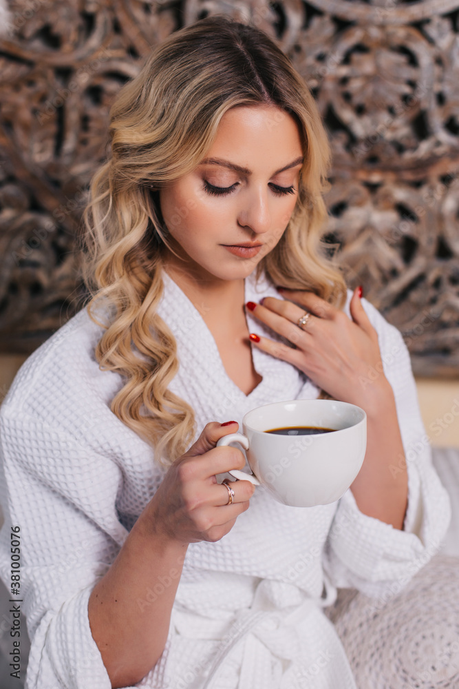 a young woman sits on a bed in the morning at home in a white bathrobe and drinks coffee. quarantined girl woke up late in the morning a young woman sits on a bed in the morning at home in a white bathrobe and drinks coffee. quarantined girl woke up late in the morning