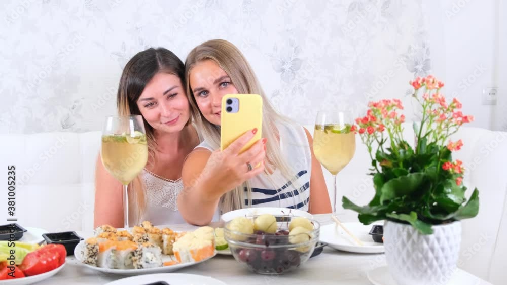 friendship and party concept - two young beautiful girlfriends drink wine and take selfie on smartphone in a cozy kitchen at the table, selective focus friendship and party concept - two young beautiful girlfriends drink wine and take selfie on smartphone in a cozy kitchen at the table, selective focus