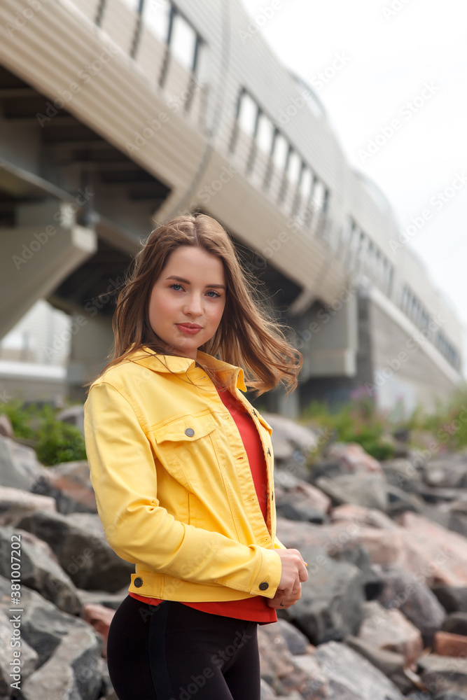 Pretty young sexy woman of athletic build is standing on stones on city beach. Walk through city center. Portrait of cute female in yellow jacket and black trousers on street