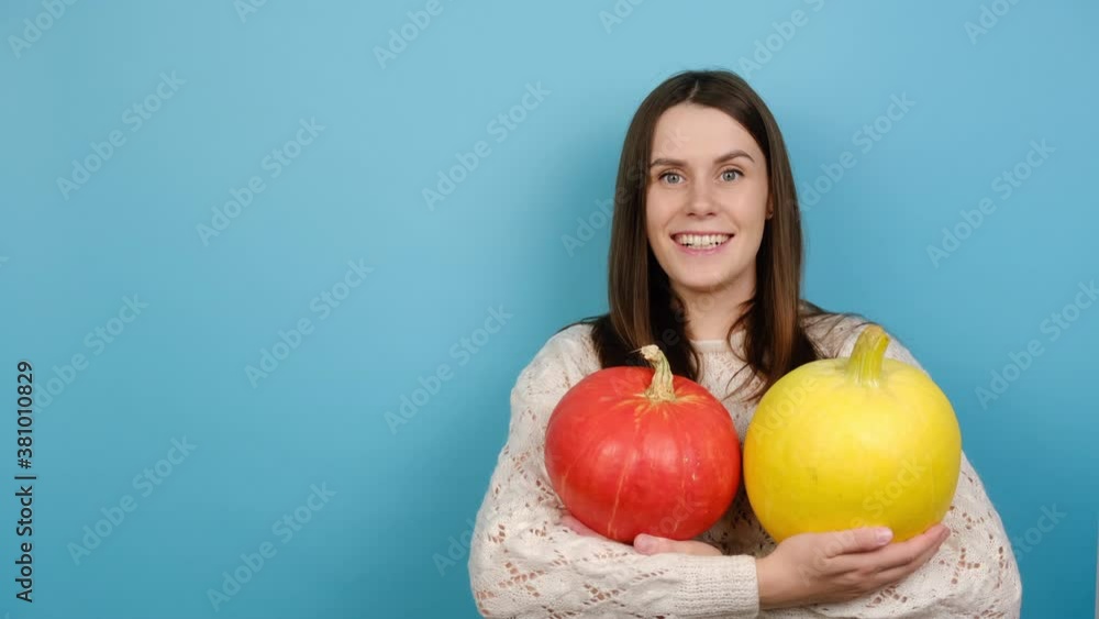 Portrait of smiling millennial woman holds two small pumpkins picked from autumn garden, wears white knitted sweater, looks in bewilderment, isolated on blue background with copy space. Hello autumn Portrait of smiling millennial woman holds two small pumpkins picked from autumn garden, wears white knitted sweater, looks in bewilderment, isolated on blue background with copy space. Hello autumn