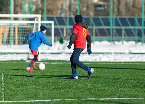 Boys in red sportswear running on soccer field with snow on background. Young footballers dribble and kick football ball in game. Training, active lifestyle, sport, children winter activity © Natali