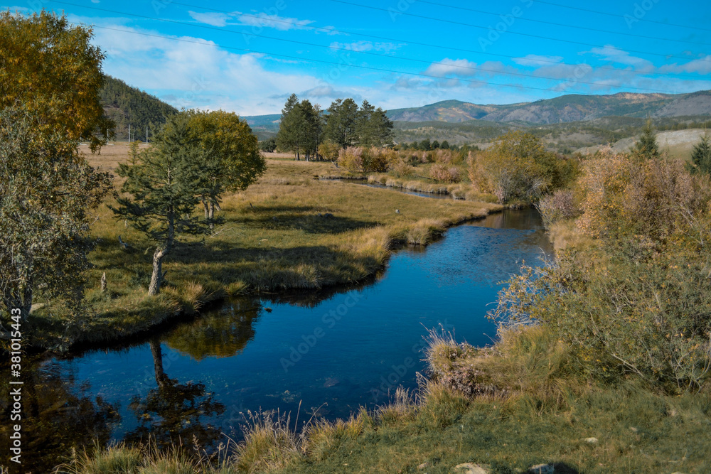 Bright blue winding river flows in grass, bushes, trees among the mountains and sky with clouds. Reflections in clear water. Autumn russia landscape