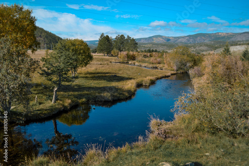 Bright blue winding river flows in grass, bushes, trees among the mountains and sky with clouds. Reflections in clear water. Autumn russia landscape