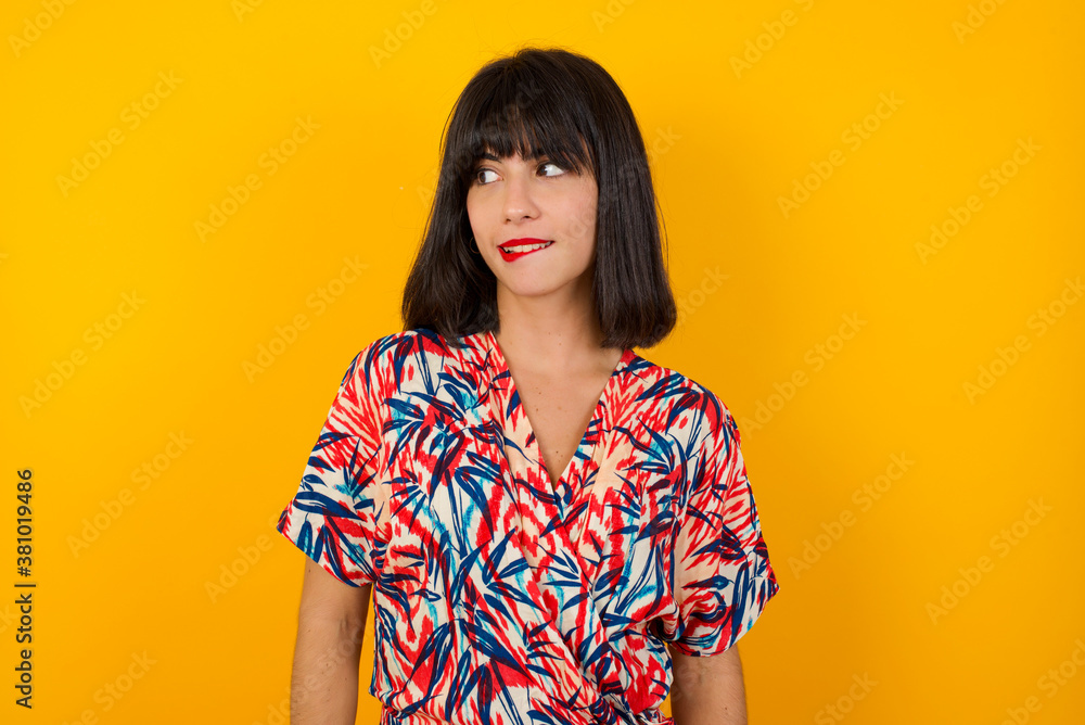 Dreamy young female with thoughtful expression, looks away keeps hands down bitting her lip thinks about something pleasant poses against gray wall. Dreamy young female with thoughtful expression, looks away keeps hands down bitting her lip thinks about something pleasant poses against gray wall.
