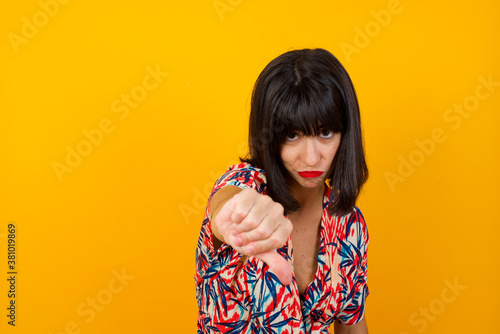 Discontent European woman shows disapproval sign  keeps thumb down  expresses dislike  frowns face in discontent  dressed in white shirt  isolated over gray background. Body language concept.