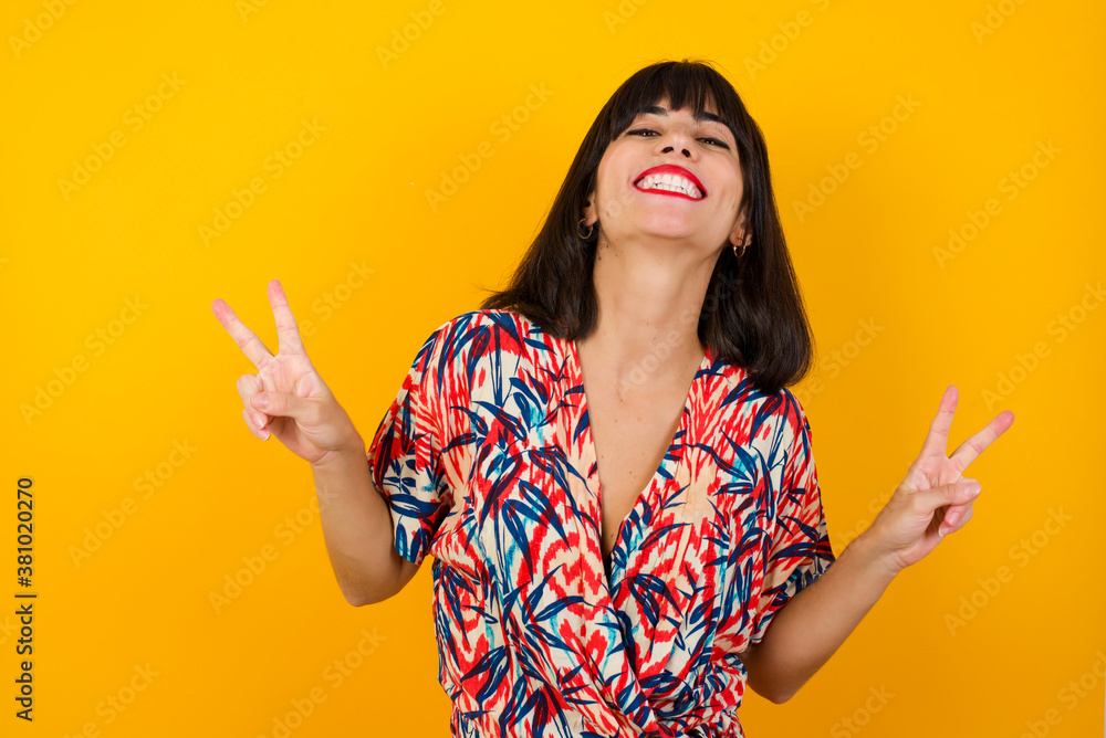 Isolated shot of cheerful woman makes peace or victory sign with both hands, dressed in casual clothes, feels cool has toothy smile, isolated over gray background. People and body language. Isolated shot of cheerful woman makes peace or victory sign with both hands, dressed in casual clothes, feels cool has toothy smile, isolated over gray background. People and body language.
