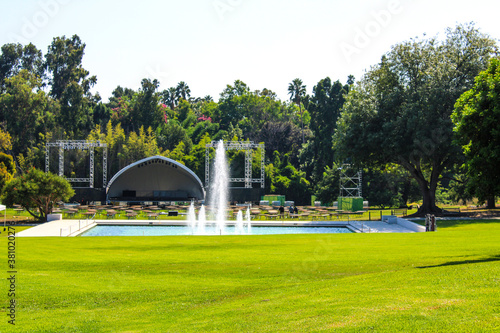 a vast stretch of lush green grass with a waterfall, lush green trees and concert venue with a stage and seats at the LA Arboretum in Arcadia California USA