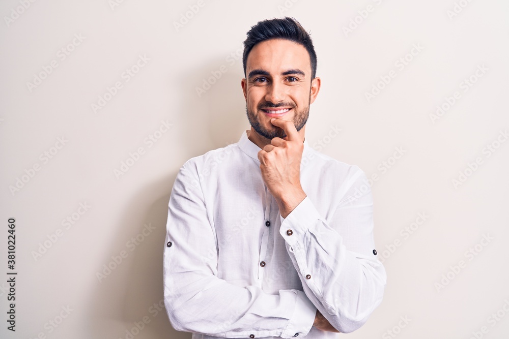 Young handsome man with beard wearing casual t-shirt standing over white background smiling looking confident at the camera with crossed arms and hand on chin. Thinking positive. Young handsome man with beard wearing casual t-shirt standing over white background smiling looking confident at the camera with crossed arms and hand on chin. Thinking positive.