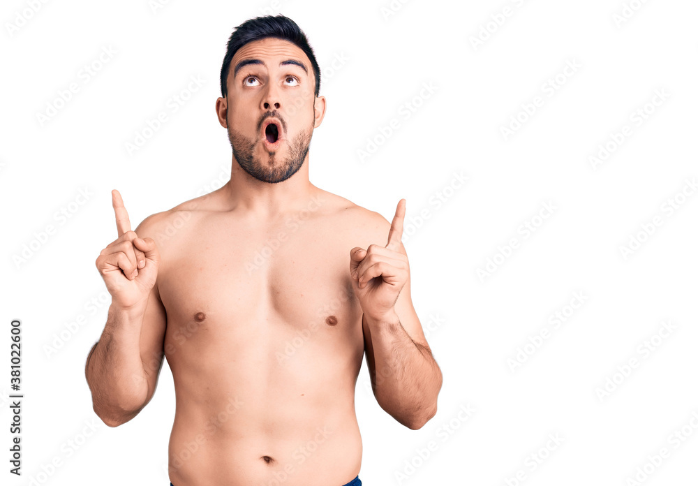 Young handsome man wearing swimwear amazed and surprised looking up and pointing with fingers and raised arms. Young handsome man wearing swimwear amazed and surprised looking up and pointing with fingers and raised arms.