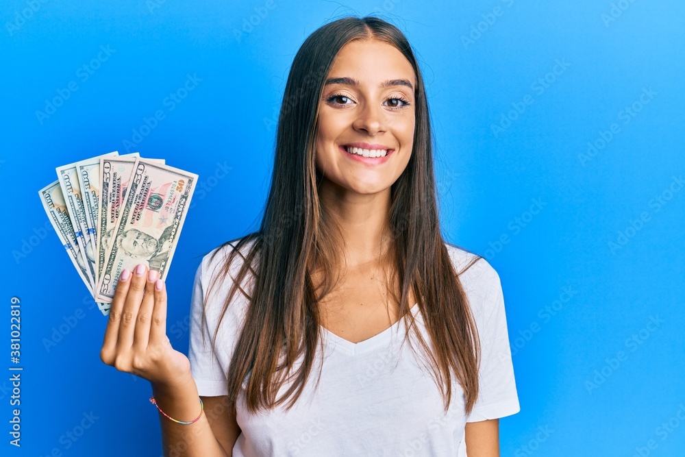 Young hispanic woman holding dollars looking positive and happy standing and smiling with a confident smile showing teeth Young hispanic woman holding dollars looking positive and happy standing and smiling with a confident smile showing teeth