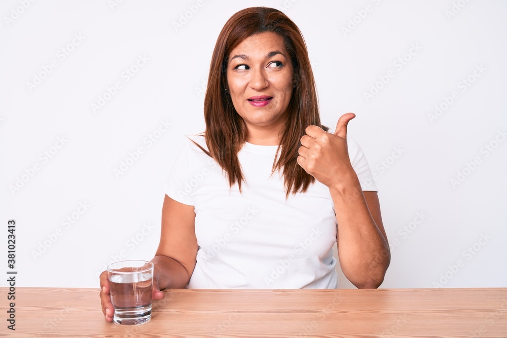 Middle age brunette hispanic woman drinking glass of water sitting on the table pointing thumb up to the side smiling happy with open mouth Middle age brunette hispanic woman drinking glass of water sitting on the table pointing thumb up to the side smiling happy with open mouth