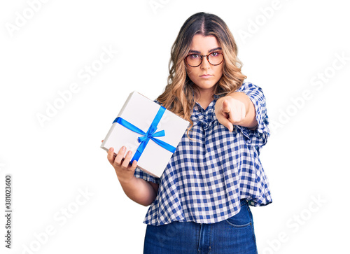 Young caucasian woman holding gift pointing with finger to the camera and to you, confident gesture looking serious