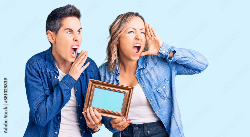 Couple of women holding empty frame shouting and screaming loud to side with hand on mouth. communication concept. Couple of women holding empty frame shouting and screaming loud to side with hand on mouth. communication concept.