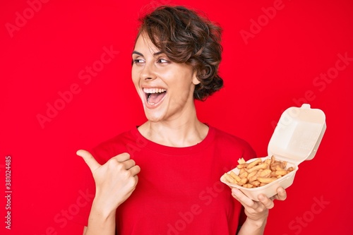 Young hispanic woman holding potato chip pointing thumb up to the side smiling happy with open mouth