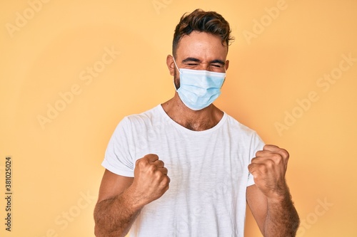 Young hispanic man wearing medical mask celebrating surprised and amazed for success with arms raised and eyes closed