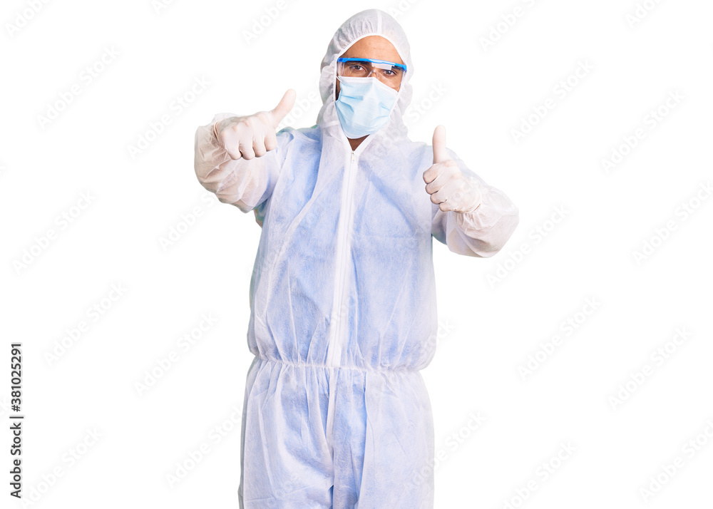 Young hispanic man wearing doctor protection coronavirus uniform and medical mask approving doing positive gesture with hand, thumbs up smiling and happy for success. winner gesture. Young hispanic man wearing doctor protection coronavirus uniform and medical mask approving doing positive gesture with hand, thumbs up smiling and happy for success. winner gesture.
