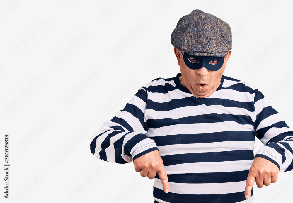 Senior handsome man wearing burglar mask and t-shirt pointing down with fingers showing advertisement, surprised face and open mouth Senior handsome man wearing burglar mask and t-shirt pointing down with fingers showing advertisement, surprised face and open mouth