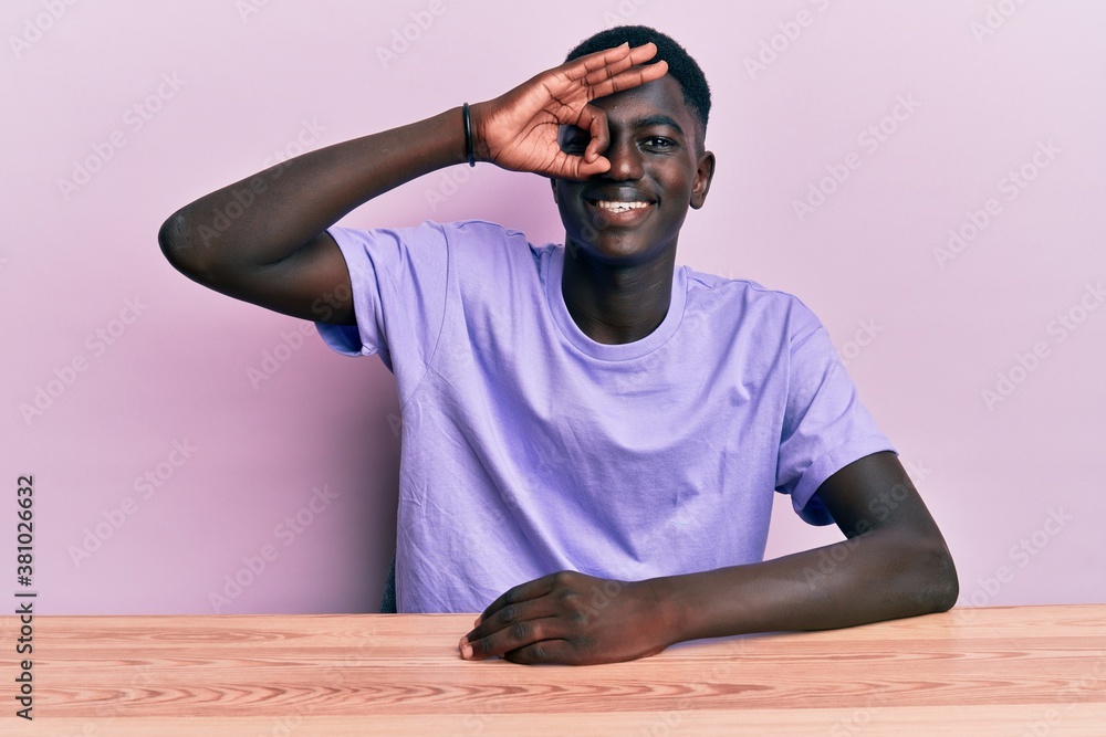 Young african american man wearing casual clothes sitting on the table smiling happy doing ok sign with hand on eye looking through fingers Young african american man wearing casual clothes sitting on the table smiling happy doing ok sign with hand on eye looking through fingers