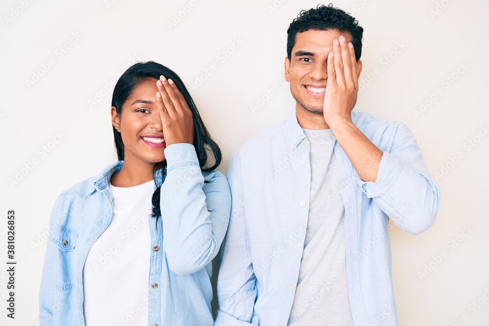Beautiful latin young couple wearing casual clothes covering one eye with hand, confident smile on face and surprise emotion.