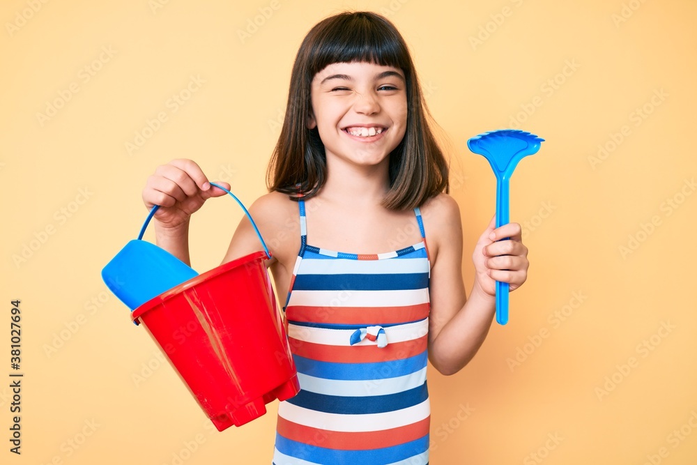 Young little girl with bang playing with summer shovel and bucket toys winking looking at the camera with sexy expression, cheerful and happy face. Young little girl with bang playing with summer shovel and bucket toys winking looking at the camera with sexy expression, cheerful and happy face.