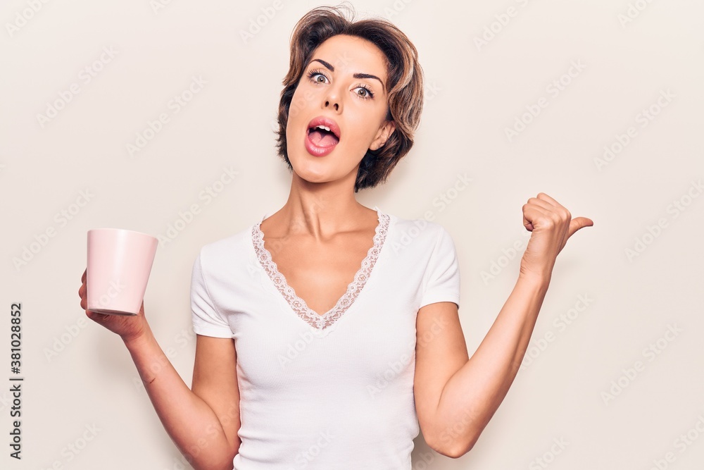 Young beautiful woman holding coffee pointing thumb up to the side smiling happy with open mouth Young beautiful woman holding coffee pointing thumb up to the side smiling happy with open mouth