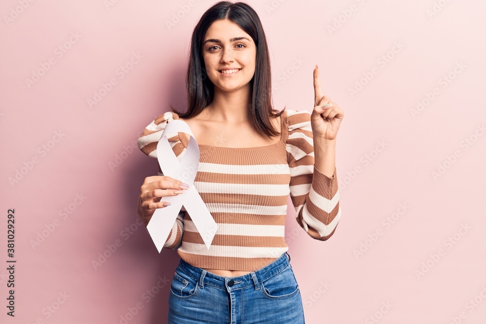 Young beautiful girl holding white cancer ribbon smiling with an idea or question pointing finger with happy face, number one Young beautiful girl holding white cancer ribbon smiling with an idea or question pointing finger with happy face, number one