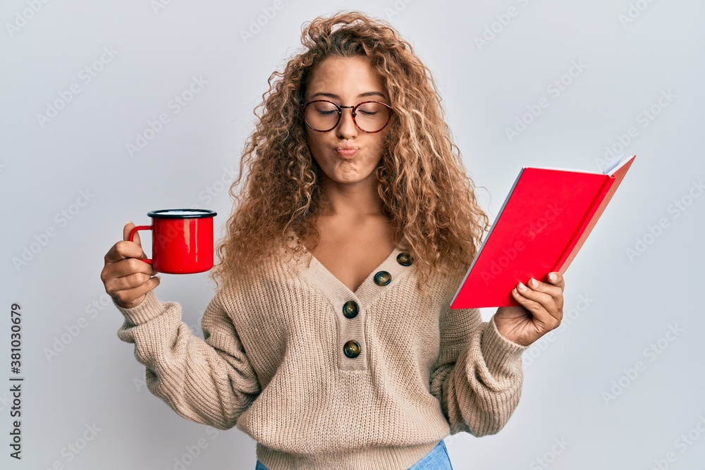 Beautiful caucasian teenager girl reading a book and drinking a cup of coffee looking at the camera blowing a kiss being lovely and sexy. love expression. Beautiful caucasian teenager girl reading a book and drinking a cup of coffee looking at the camera blowing a kiss being lovely and sexy. love expression.