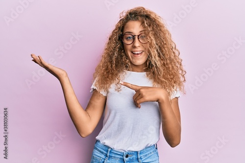 Beautiful caucasian teenager girl wearing white t-shirt over pink background amazed and smiling to the camera while presenting with hand and pointing with finger.