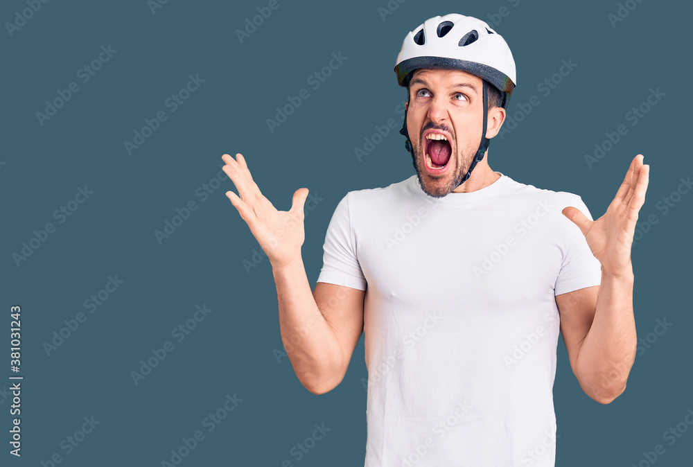 Young handsome man wearing bike helmet crazy and mad shouting and yelling with aggressive expression and arms raised. frustration concept. Young handsome man wearing bike helmet crazy and mad shouting and yelling with aggressive expression and arms raised. frustration concept.