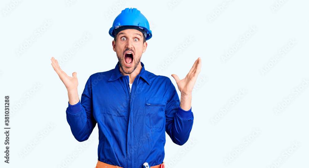 Young handsome man wearing worker uniform and hardhat celebrating crazy and amazed for success with arms raised and open eyes screaming excited. winner concept