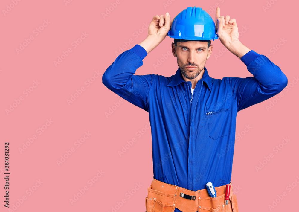 Young handsome man wearing worker uniform and hardhat doing funny gesture with finger over head as bull horns Young handsome man wearing worker uniform and hardhat doing funny gesture with finger over head as bull horns