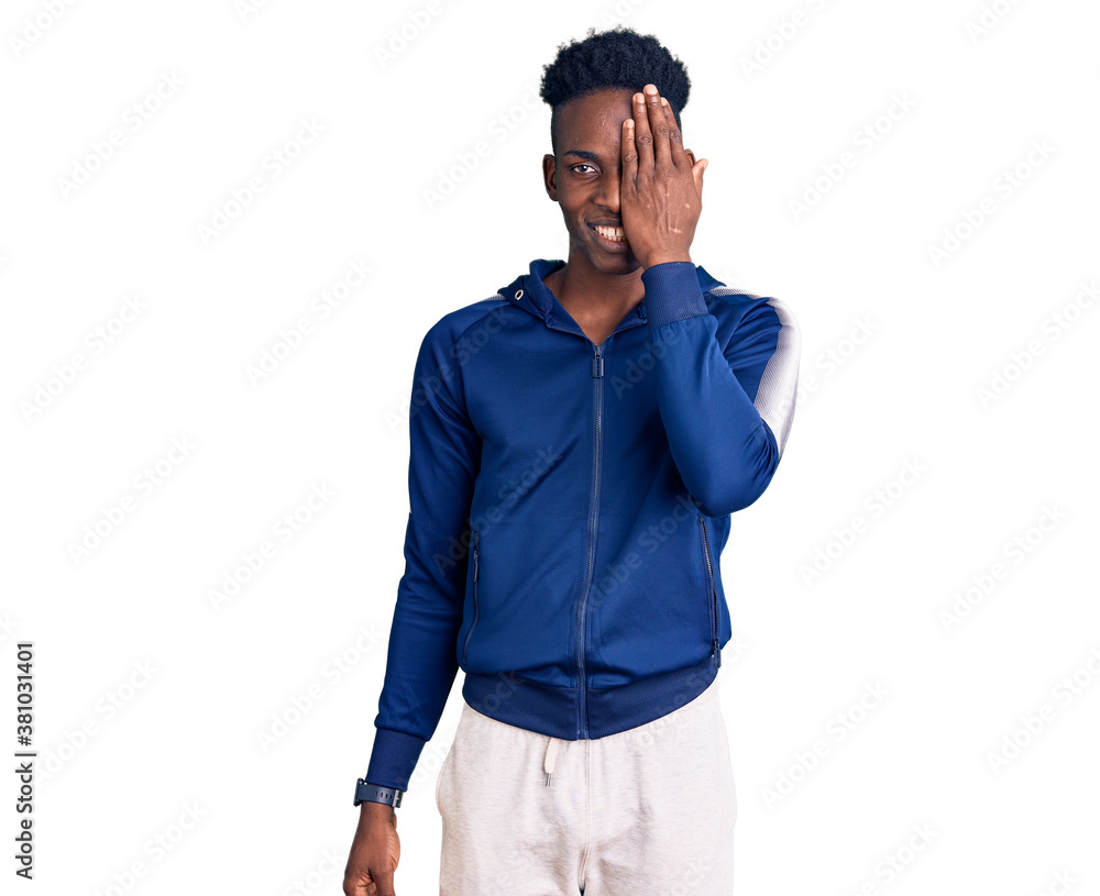 Young african american man wearing sportswear covering one eye with hand, confident smile on face and surprise emotion. Young african american man wearing sportswear covering one eye with hand, confident smile on face and surprise emotion.