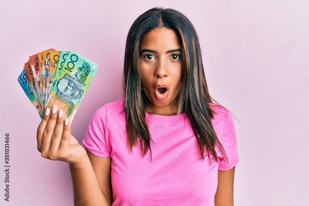 Young latin girl holding australian dollars scared and amazed with open mouth for surprise, disbelief face Young latin girl holding australian dollars scared and amazed with open mouth for surprise, disbelief face