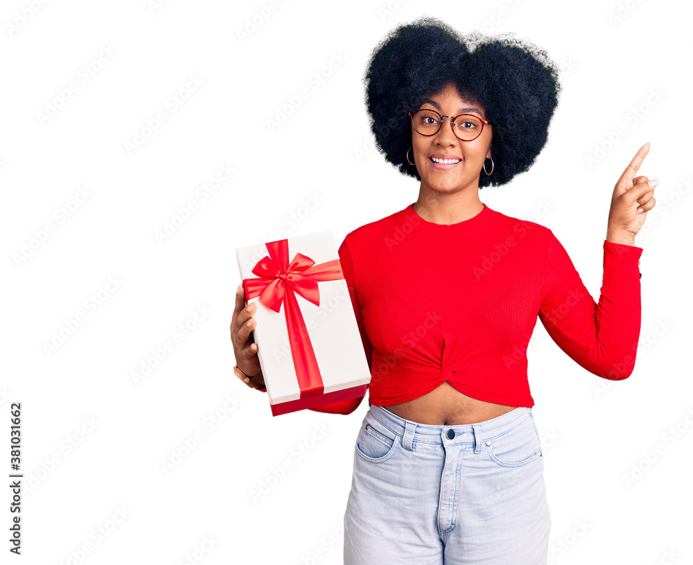Young african american girl holding gift with a big smile on face, pointing with hand finger to the side looking at the camera. Young african american girl holding gift with a big smile on face, pointing with hand finger to the side looking at the camera.