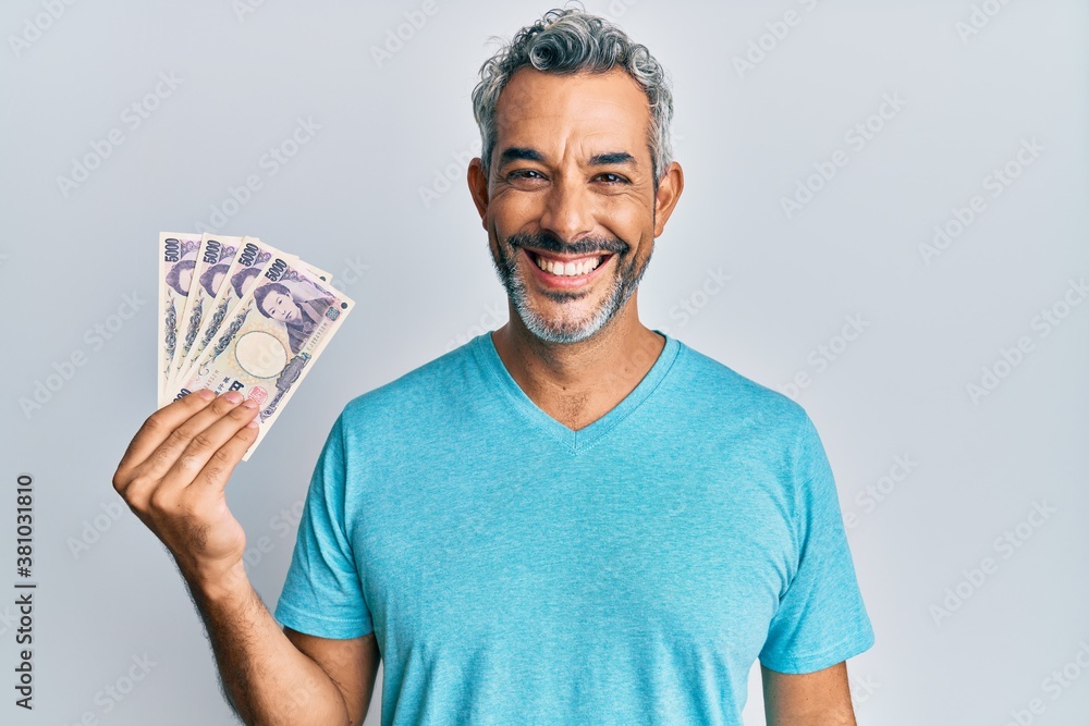 Middle age grey-haired man holding japanese yen banknotes looking positive and happy standing and smiling with a confident smile showing teeth Middle age grey-haired man holding japanese yen banknotes looking positive and happy standing and smiling with a confident smile showing teeth