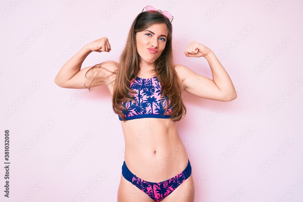 Obraz premium Beautiful young caucasian woman wearing bikini and sunglasses showing arms muscles smiling proud. fitness concept.