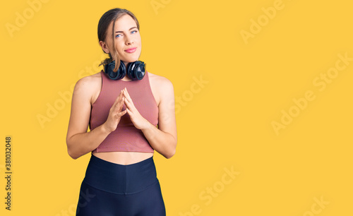 Beautiful caucasian young woman wearing gym clothes and using headphones hands together and fingers crossed smiling relaxed and cheerful. success and optimistic