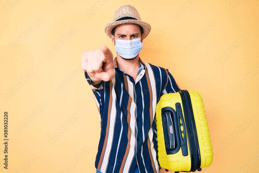 Young handsome man wearing medical mask holding suitcase pointing with finger to the camera and to you, confident gesture looking serious