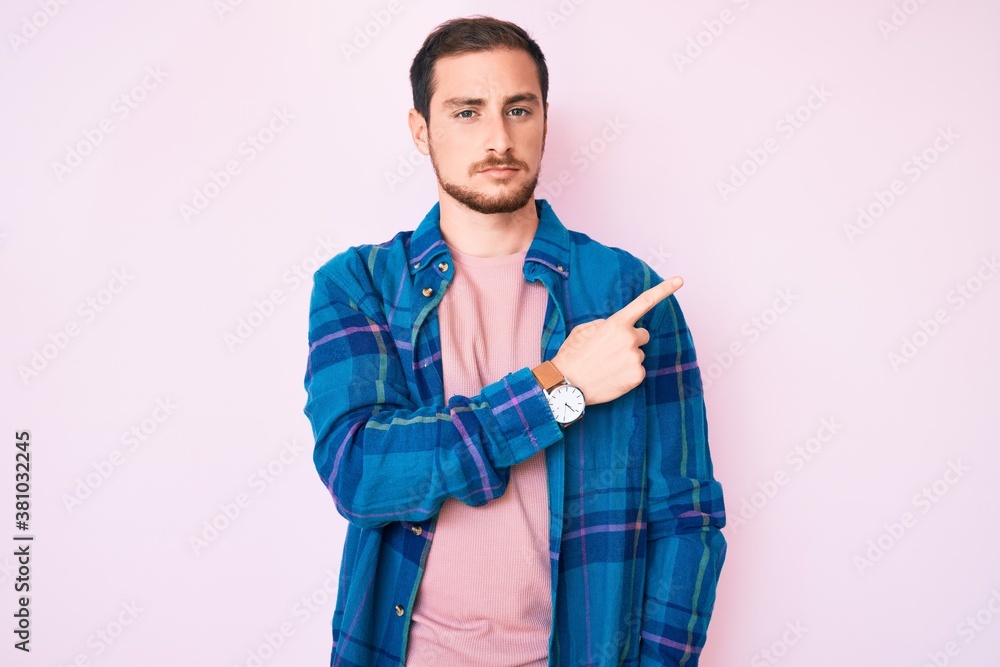 Young handsome man wearing casual clothes pointing with hand finger to the side showing advertisement, serious and calm face Young handsome man wearing casual clothes pointing with hand finger to the side showing advertisement, serious and calm face