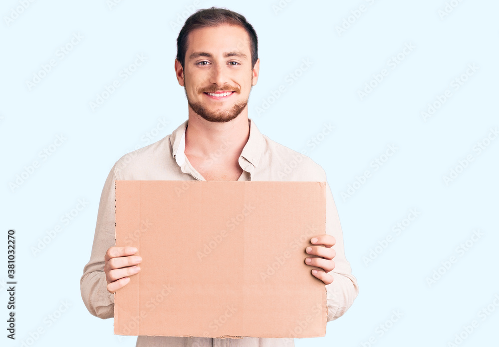 Young handsome caucasian man holding empty cardboard banner looking positive and happy standing and smiling with a confident smile showing teeth Young handsome caucasian man holding empty cardboard banner looking positive and happy standing and smiling with a confident smile showing teeth
